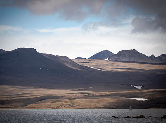 Rugged Icelandic landscape with mountains, snow patches, and a body of water under an overcast sky.