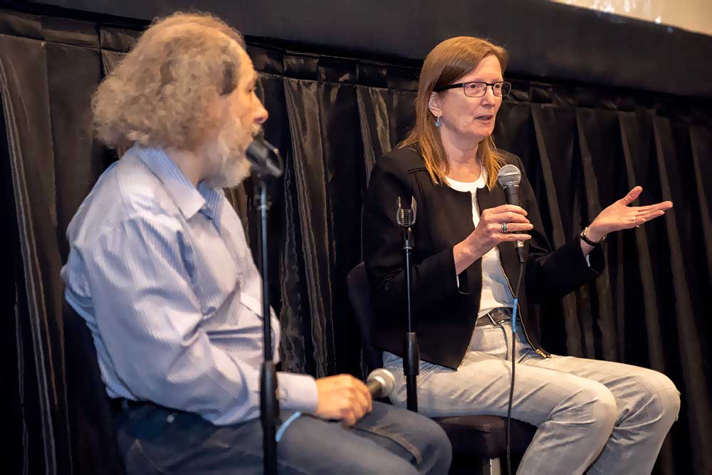 photo of two seated hosts speaking on stage