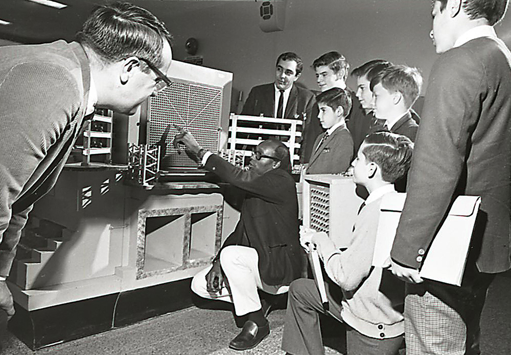 Black and white photo of Gus Prince surrounded by students as he points to reactor equipment