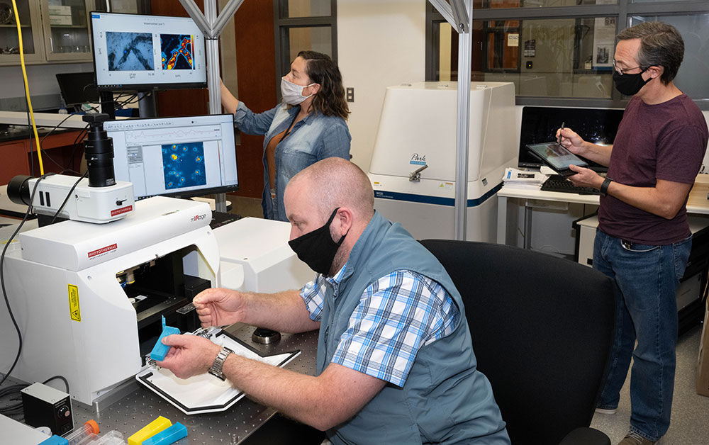 Photo of Samuel Tenney (sitting front), Sabine Neal (standing near screens), and Dario Stacchiola at