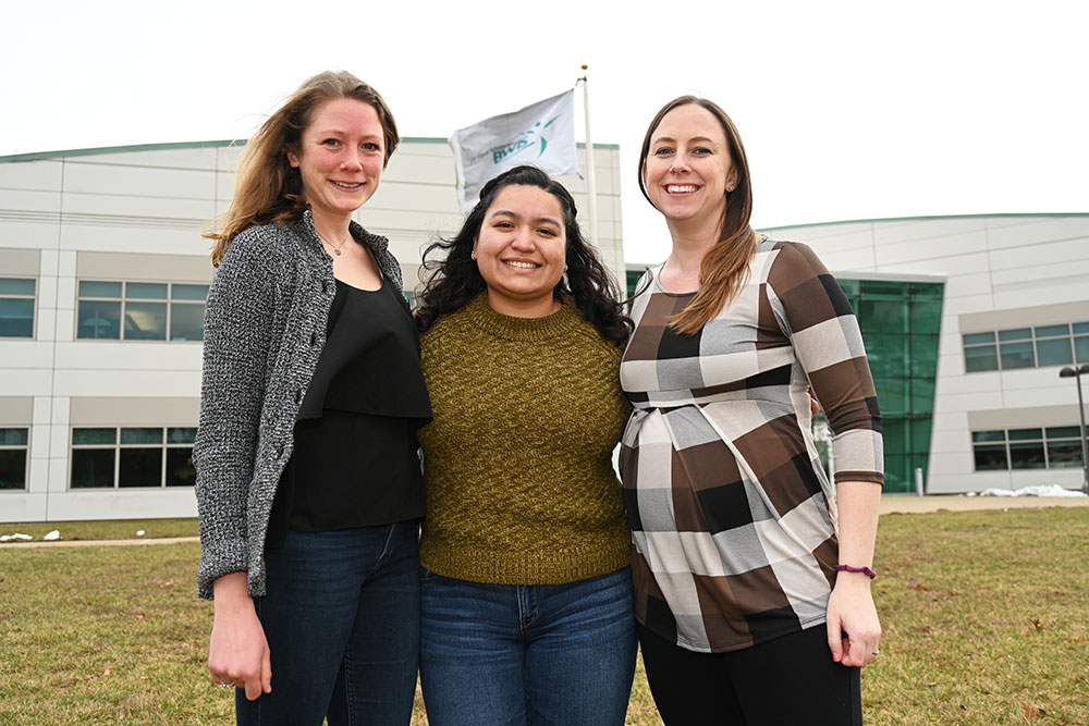 Jessica Gasparik, Jennefer Maldonado, and Megan Magrum pose in front of a flag pole flying the BWIS
