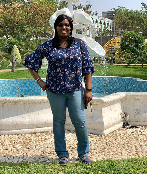 Ogo, wearing sunglasses, blue blouse and jeans, stands in front of a white stone fountain