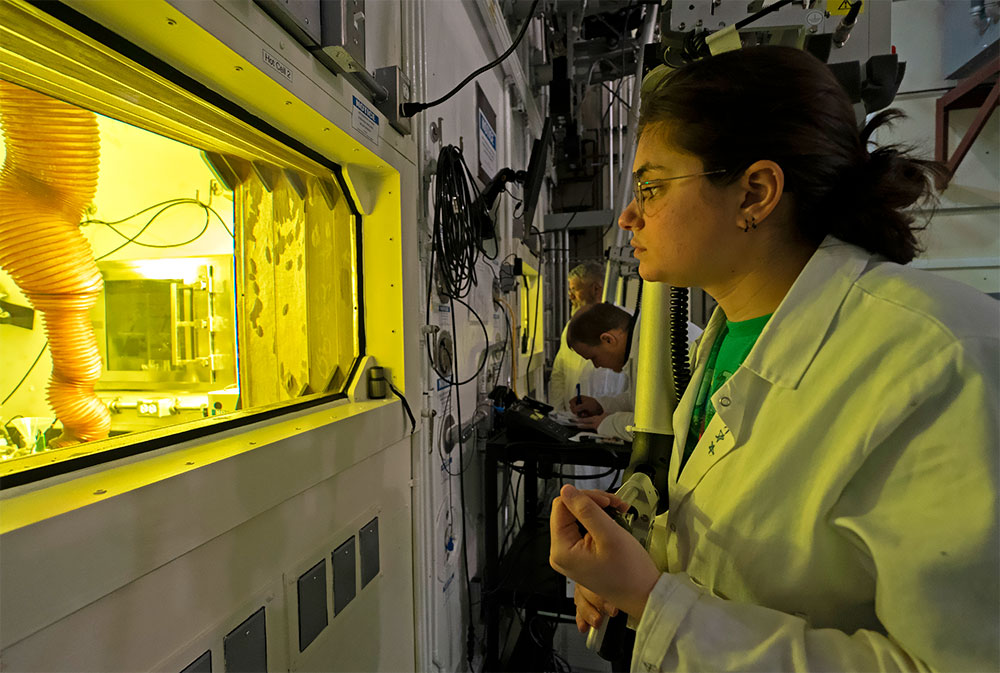 An MIRP team member, wearing glasses and a white lab coat, looks a hot-cell window that has a yellow