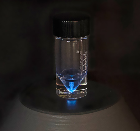 A vial surrounded by darkness shows a small amount of liquid that glows blue via long exposure photo