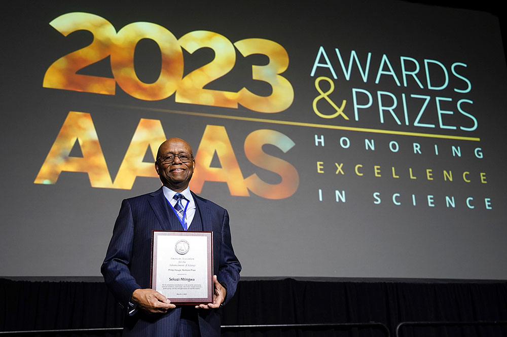 Sekazi Mtingwa holds an award plaque in front of a project banner that reads "2023 AAAS Awards