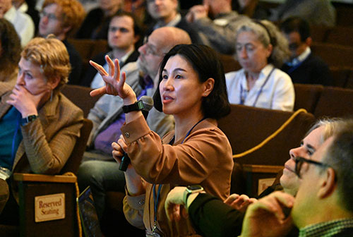 A woman seated in an auditorium speaks into a microphone she is holding