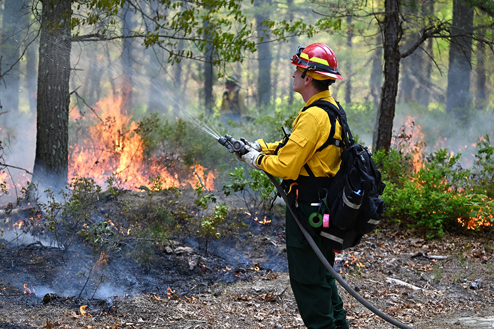 Firefighter wearing a yellow jacket, red hat uses a hose to spray water on a fire