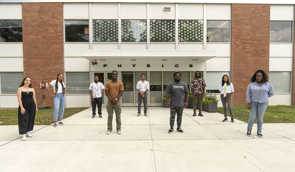 Nine students stand in a line in front of physics building