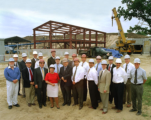 A group of people wearing hardhats pose for a photo with a building frame and construction equipment