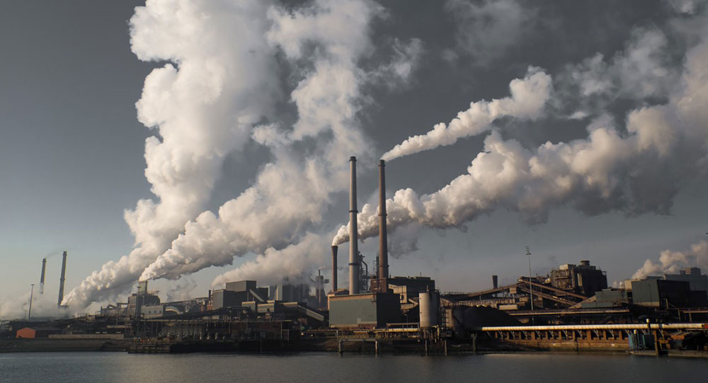 Clouds of emissions rise from smokestacks from a waterfront industrial complex.