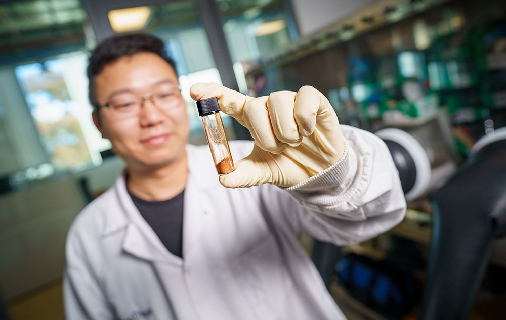 Scientist in a white lab coat with a gloved hand holds a vial containing brown powder.