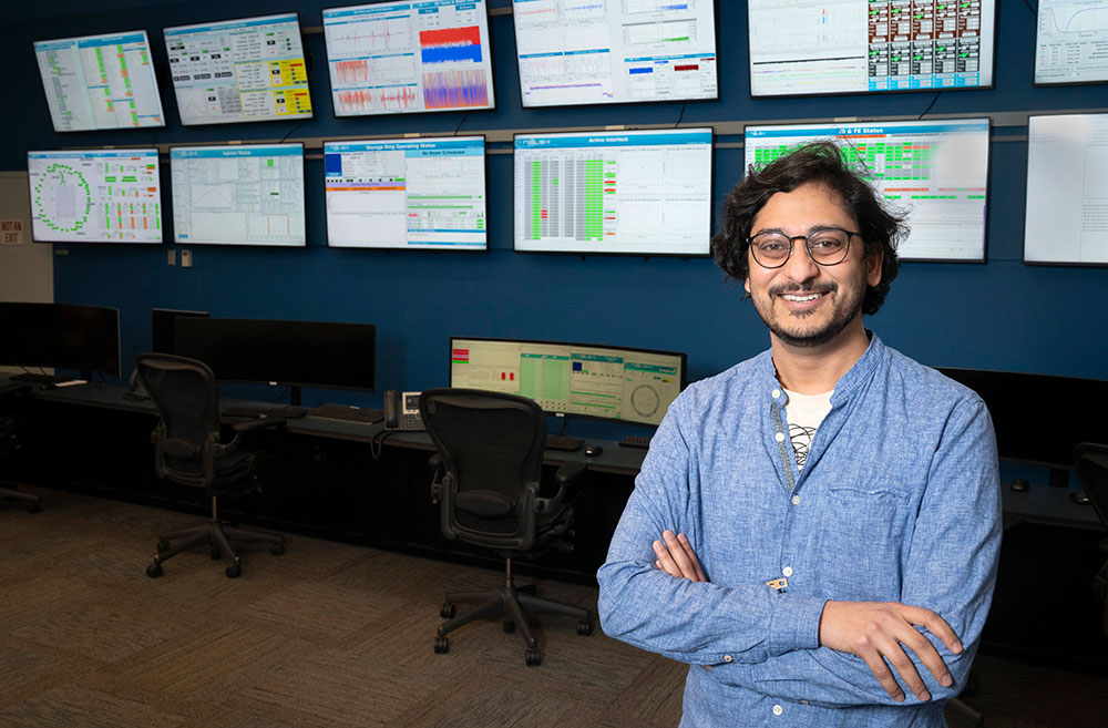 A man wearing glasses and a blue collared shirt smiles with multiple computer screens in the backgro