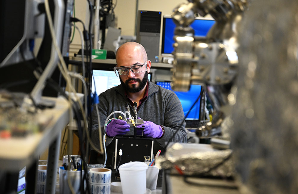 A bearded man in a gray jumper with a red collar wears glasses and safety goggles. He looks down as