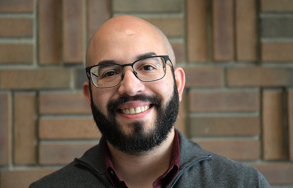 A bearded man wearing glasses smiles at the camera. He wears a gray jumper with a red collar. The wa