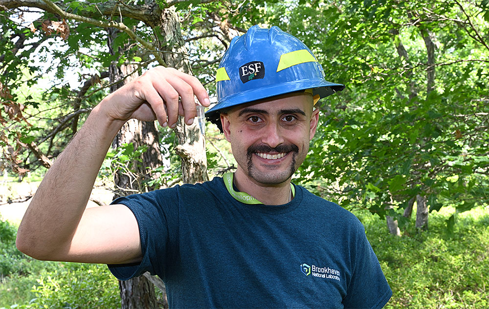 A man in a blue hard hat grins into the camera, center-frame. He sports a handlebar moustache and we
