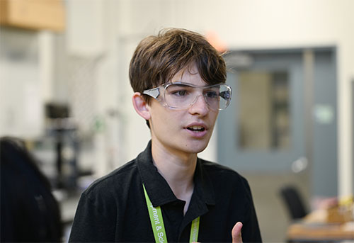 A young man in safety goggles and a collared, black shirt with a neon green lanyard around his neck