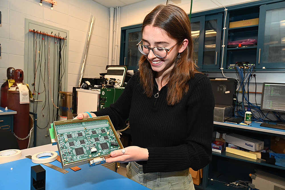 A young woman in glasses and a black pullover looks down at a rectangular board of circuitry in hues