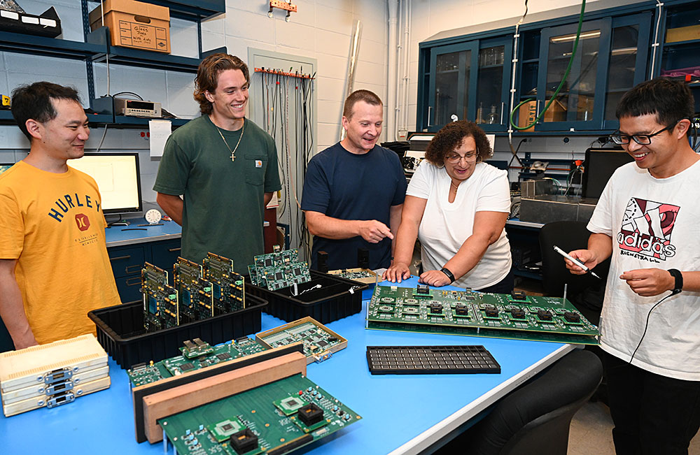 Five individuals gather in a semi-circle around a blue workbench laden with electronic circuit board