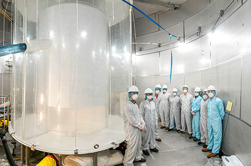 At left, a clear, large cylindrical water tank. Right: A group of nine team members in personal prot