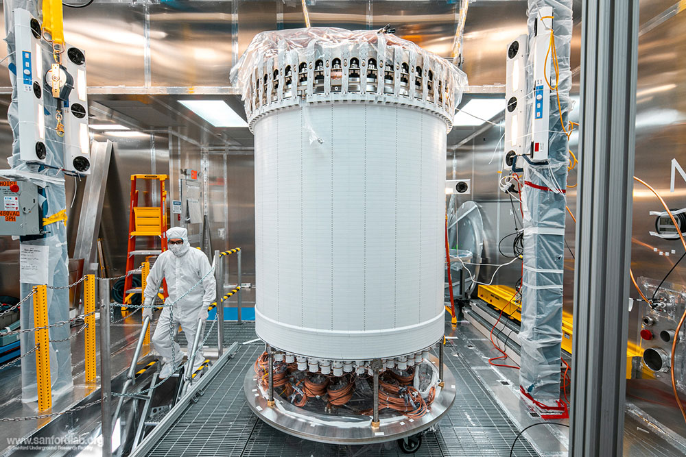 A large, white cylindrical detector at the center of a lab. At left, a person in head-to-toe white p