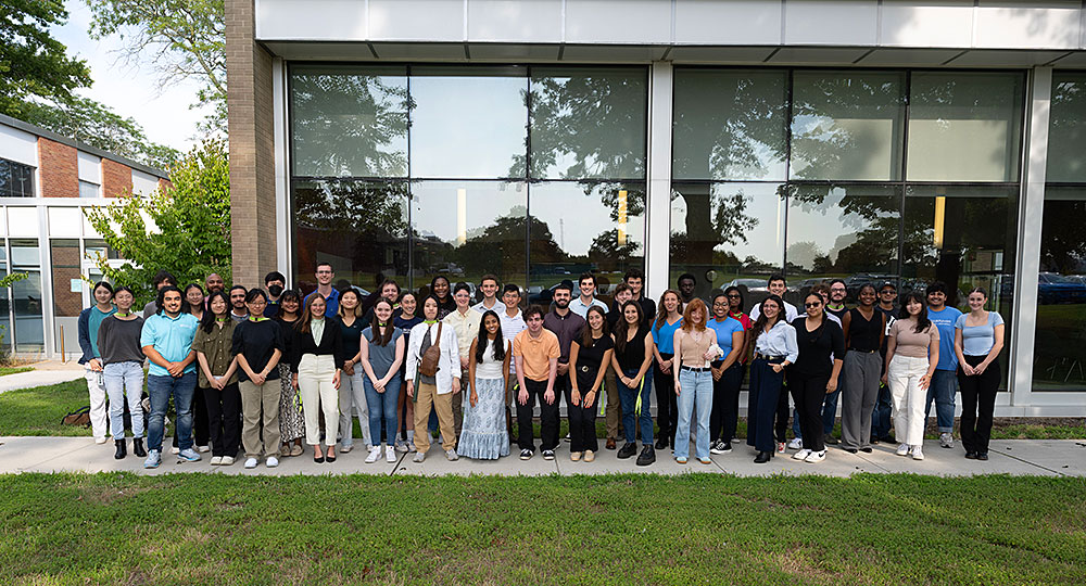 A group of interns pose together outside a building with tall glass windows