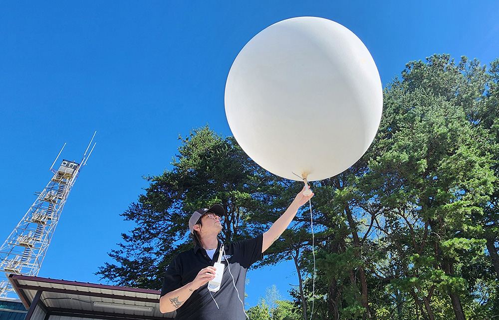 A person holds the bottom of a weather balloon with their left hand and a radiosonde in their right