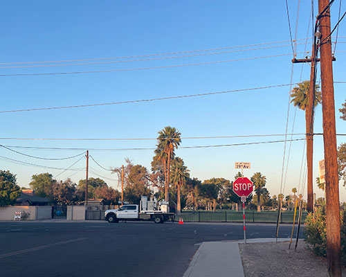 A truck with instruments is parked along a street next to a fence with grass and trees beyond it. A