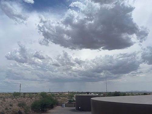 Rain clouds float above a desert landscape with some vegetation and buildings. Streaks of rain are s
