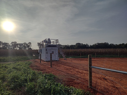 A lidar sits on top on an instrument container sited on dark red soil. Just to the left of the conta