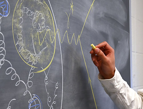 A scientist's holds a yellow piece of chalk up to a chalboard