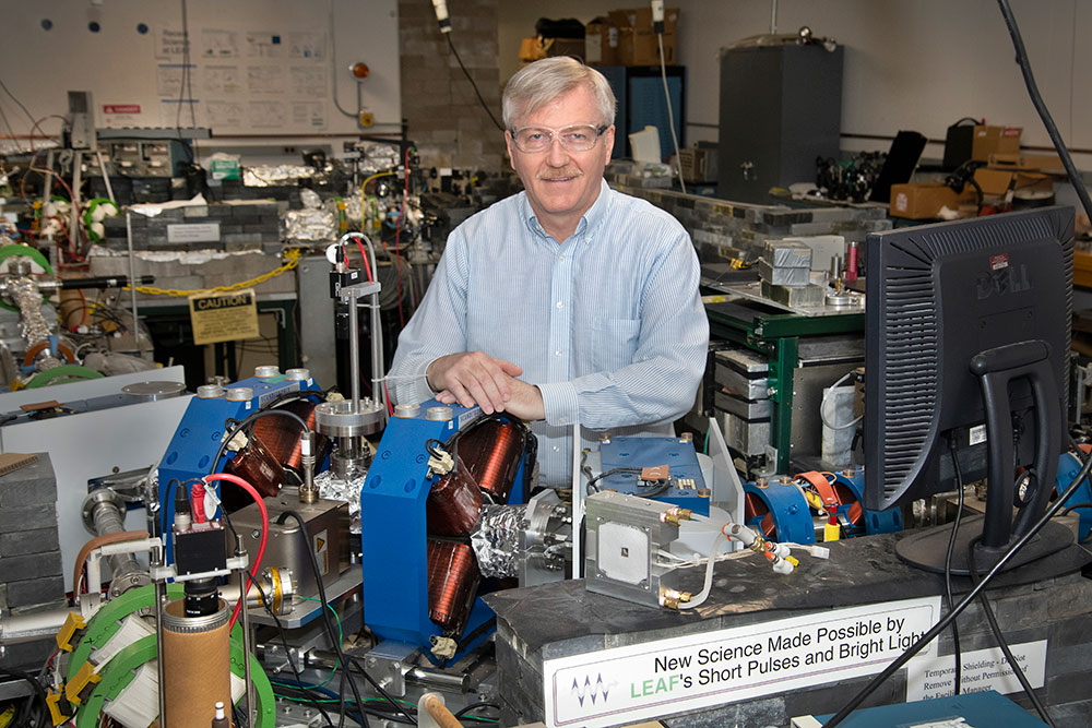 James Wishart, wearing clear safety glasses and a blue button-down, stands behind the LEAF laborator