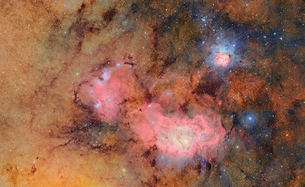 A cosmic tapestry of glowing tan and pink gas clouds with dark dust lanes. In the upper right, the T