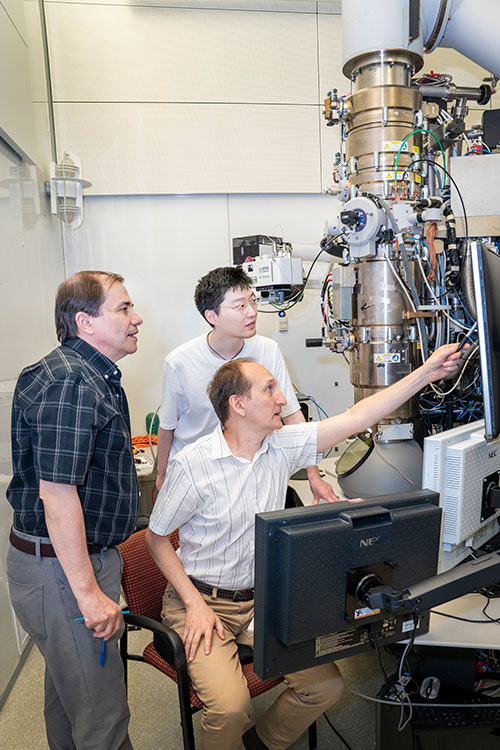 Three researchers examine a monitor next to a large microscope