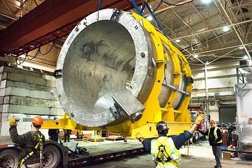 Technicians guide a large cylindrical magnet lifted inside a warehouse space