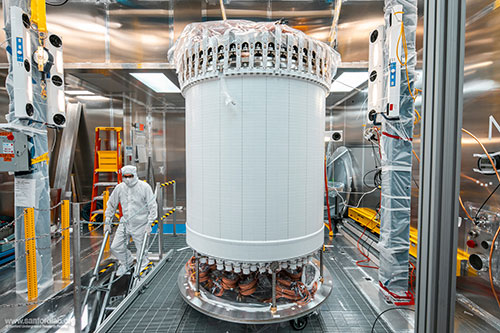 A researcher in protective gear works near a tall, cylindrical scientific instrument with visible wi