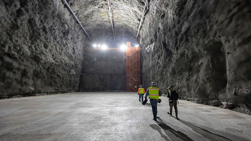 Workers in safety gear walk across a large underground cavern with high rock walls and bright overhe