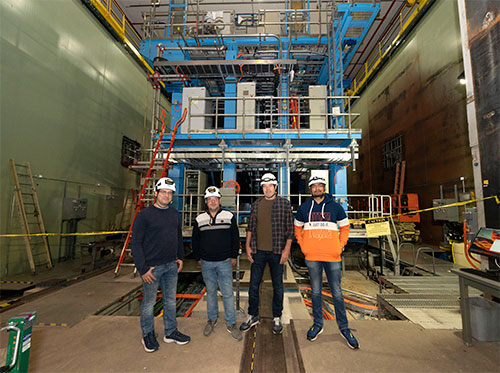Four workers wearing hard hats stand before the sPHENIX detector, with large green shielding walls v