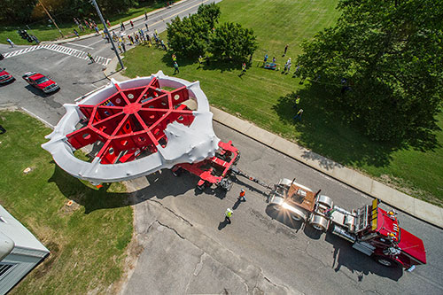 Large circular magnet is transported by truck along a road with workers and onlookers nearby