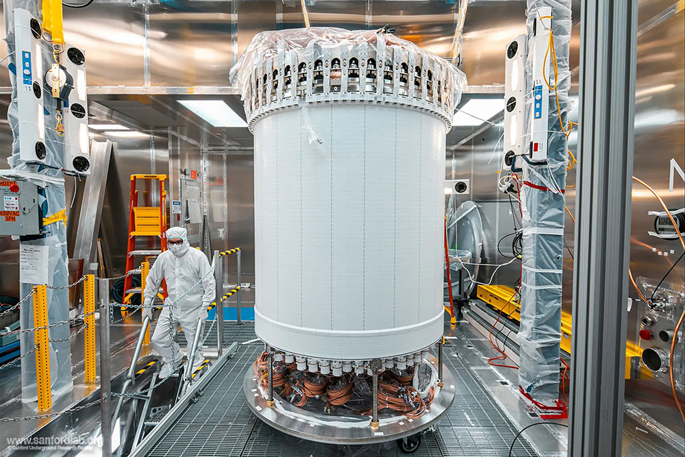 A researcher in protective gear works near a tall, cylindrical scientific instrument with visible wi