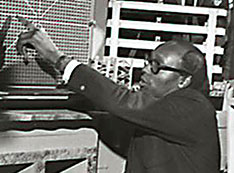 Black and white photo of Gus Prince surrounded by students as he points to reactor equipment