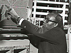 Black and white photo of Gus Prince surrounded by students as he points to reactor equipment