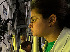 An MIRP researcher, wearing glasses and a white lab coat, looks a hot-cell window that has a yellow