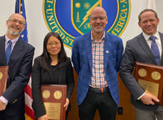 Wah-Keat Lee, Lijuan Ruan, John Hill, and Alistair Rogers pose in front of a large DOE seal