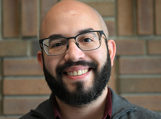 A bearded man wearing glasses smiles at the camera. He wears a gray jumper with a red collar. The wa