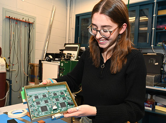 A young woman in glasses and a black pullover looks down at a rectangular board of circuitry in hues