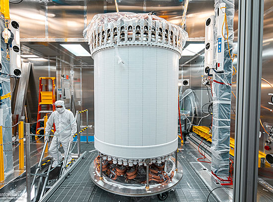 A large, white cylindrical detector at the center of a lab. At left, a person in head-to-toe white p