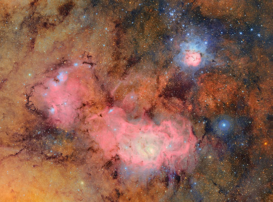 A cosmic tapestry of glowing tan and pink gas clouds with dark dust lanes. In the upper right, the T