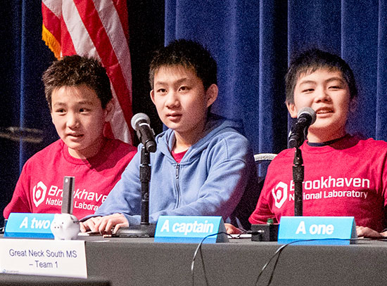 Students wearing Brookhaven National Laboratory T-shirts sit behind microphones on a stage during a