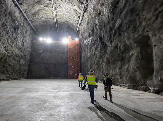 Workers in safety gear walk across a large underground cavern with high rock walls and bright overhe
