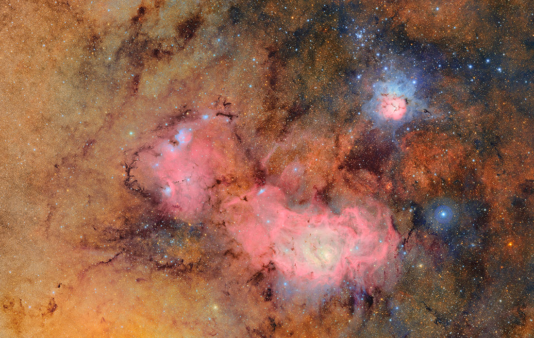 A cosmic tapestry of glowing tan and pink gas clouds with dark dust lanes. In the upper right, the T